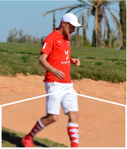 A player in a red shirt walking by a sand pit with a palm tree in the background.