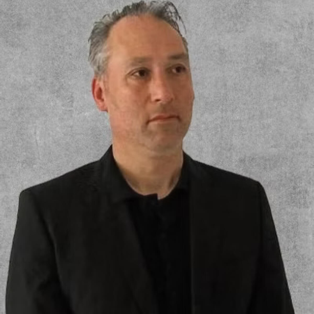 A headshot of a man in a black suit and a black shirt standing in front of a grey wall.