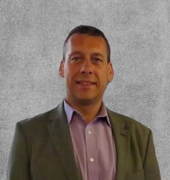 A man in a suit and purple shirt stands confidently in front of a gray wall.