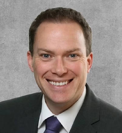 A close-up headshot of a man in a brown suit and a blue tie, smiling.