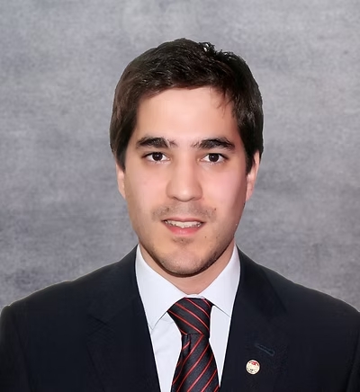 A headshot of a man in a black suit and a black and red tie, standing in front of a wall.