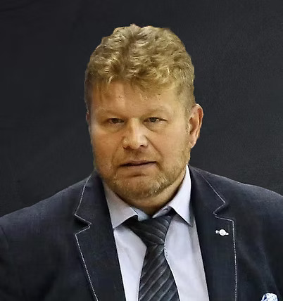 A headshot of a man in a black suit in front of a dark grey wall.