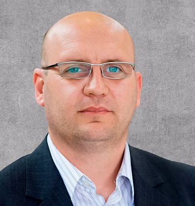 A close-up portrait photo of a man with glasses in a black suit and a blue shirt.