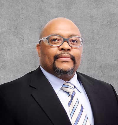 A portrait photo of a man with glasses in a black suit, blue shirt, and tie.